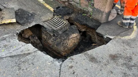 A picture of the sinkhole under the road. A hole in the road surface can be seen with workers attending the scene.