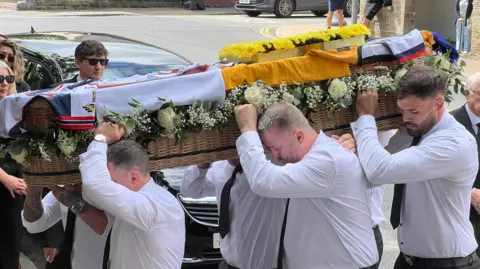 Men in long-sleeved white shirts carry a coffin dressed in rugby shirts and white roses.