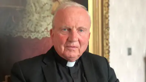 Bishop Donal McKeown is dressed in a black shirt and blazer with a white collar. 
He is sitting down in front of a large painting within a golden frame. 