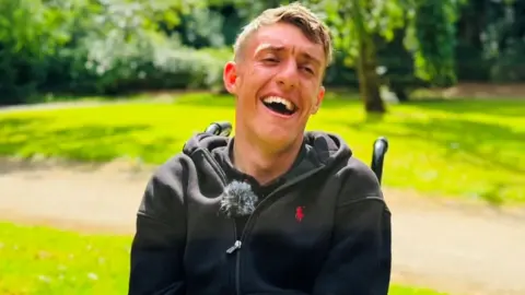 Jake Smith, a young man in a wheelchair, he is laughing heartily in a green park setting.