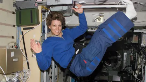 NASA Eileen Collins in the air on a spaceship. She smiles and waves at the camera, her legs raised in the air. She is wearing a blue top and pants with white socks. She holds onto a bar with her left hand and is surrounded by wires and electronic equipment.