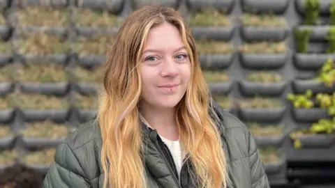 A young woman with long blonde hair wearing a green jacket and white t-shirt.