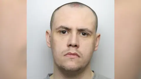 West Yorkshire Police A police custody picture of Adam Martin. He has a shaved head and stubble and is wearing a grey jumper