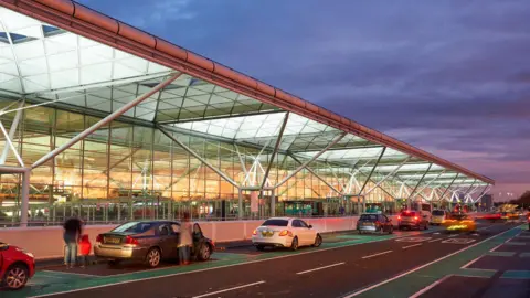 A steel and glass airport terminal building with cars parked in front of it. It is dusk and the sky is purple.