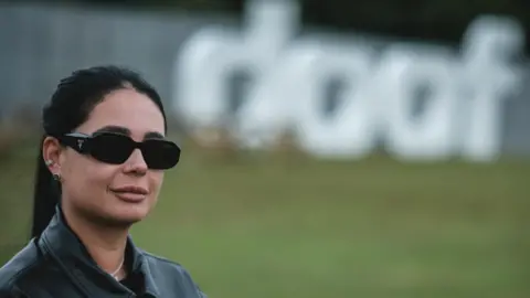 Hannah Laing, in dark sunglasses, her dark hair in a ponytail and wearing a black leather jacket, stands with a large "Doof" sign behind her in Camperdown Park