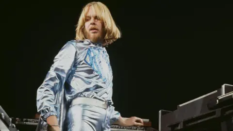 Rick Wakeman during a concert in the 1970s. He is turning away from a keyboard as he rests one hand near it. He is wearing a light blue outfit.