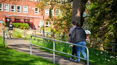 UHI Perth Students walk through the campus at UHI Perth. There is a red brick building and a path lined by trees.