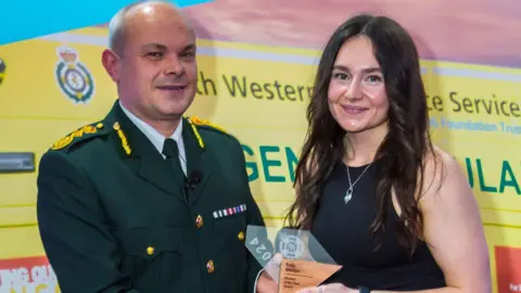 A man from South Western Ambulance Service Foundation NHS Trust with Emily Bracken. He is wearing his uniform, which includes a white shirt, a green tie and a green jacket with gold details. Emily is standing next to him. She has long, dark, curly hair and is wearing a black top and silver necklace. She is holding a Student of the Year award. Both of them are looking at the camera and smiling.