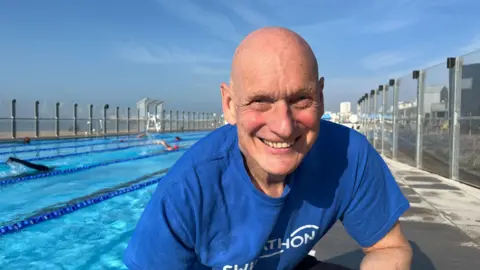 Duncan Goodhew wearing a blue Swimathon t-shirt and kneeling by the outdoor pool at sea lanes on Brighton seafront