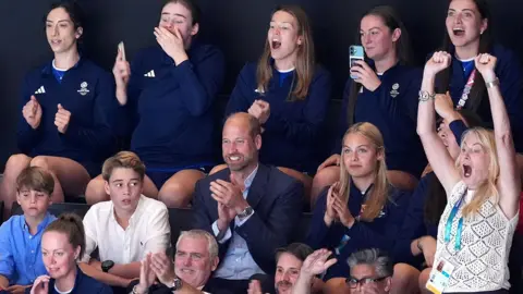 The Duke and Duchess of Rothesay with their three children watching a netball match. Everyone is seated while cheering, apart from a blonde woman on the far right of the photo who is standing with her arms in the air. She appears to be cheering loudly.