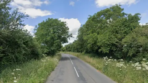 Google A rural road with one lane of traffic running in either direction. The road has long grass, hedgerows and trees on either side of it. 