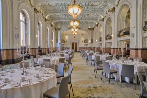 Grand Hotel Image shows a large room with an ornate ceiling and around a dozen round tables, covered in white tablecloths, grey chairs 