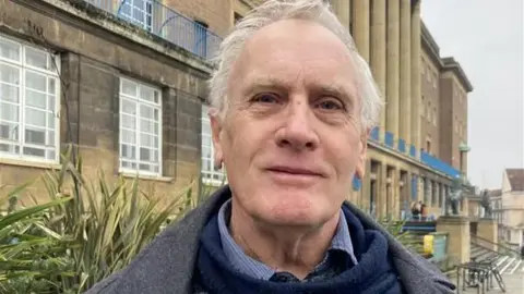 LDRS Councillor Mike Sands stood outside Norwich City Hall. He has short grey hair, is cleanly shaven and is wearing a grey jacket over a blue jumper and patterned shirt