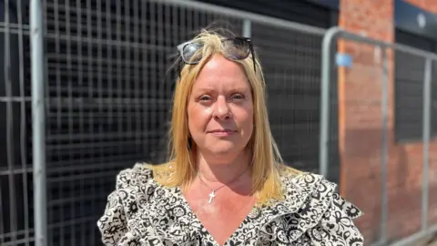 A woman with blonde straight hair and sunglasses on her head wearing a black and white summer dress stares at the camera not smiling in front of some metal railings