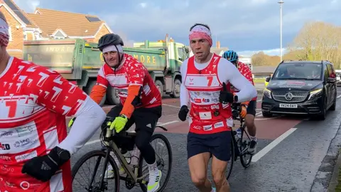 Kevin Sinfield wearing gloves and a headband running along a road, giving a thumbs-up sign as he passes a camera. On his right and behind him are people on bikes, with another runner just out of shot and a black support vehicle behind him