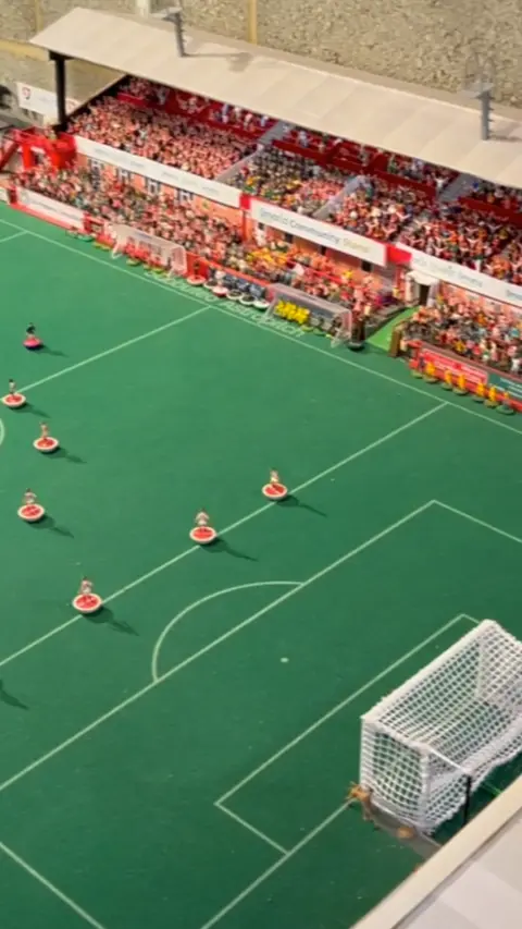 A replica model of Cheltenham Town Stadium with football players standing in the middle of the pitch and the stands full of fans.