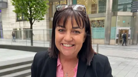 Lee-Bernadette Walford, a woman with brown hair with glasses on her head, is wearing a black suit jacket and pink blouse. She is stood outside a court building in Manchester.
