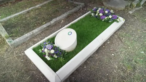 A grave with grass covering the top and a stone border. The grave has a small, circular headstone in the centre with white, purple and green flowers on top.