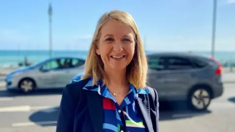 Sussex PCC a woman smiles on a sunshine day. He has a blonde shoulder length hair and a bright blue shirt. She is standing outside a busy road with cars.