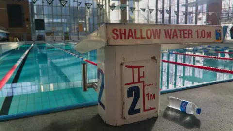 A indoor swimming pool with large glass windows behind it and a sign in the foreground which reads shallow water