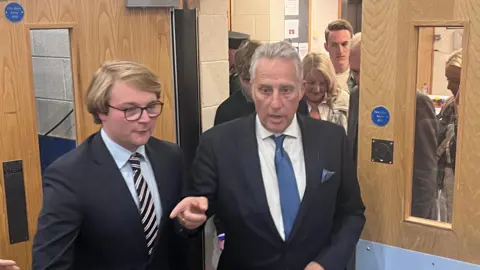 Ian Paisley arriving through a door at the count centre. He is wearing a dark suit and a blue tie and is pointing with his right hand.