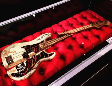 White bass guitar split in two with a skull and crossbones and pressure stickers stuck on it. It is presented on a large red cushion in a glass case