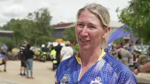 Lorna Clarke with blond hair tied back. She is looking to the left of the camera. She is wearing a blue top with a Northamptonshire Police logo. There are people behind her and what appears to be a blue inflatable slide. There are trees to the left and right.