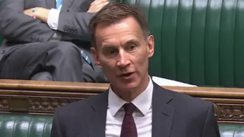 MP Jeremy Hunt wearing a grey suit jacket, white shirt and red tie, while stood asking a question in parliament