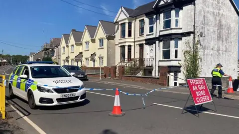 A police car is seen along a road, with police tape and a red "Road Closed" sign