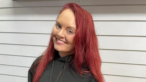 Jennie Doolan is smiling directly at the camera. She is wearing a black hooded jumper and is standing in front of a white panelled wall.