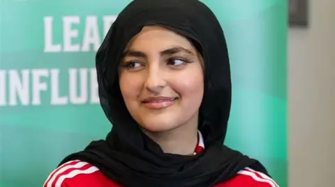 Eleeza Khan Eleeza Khan pictured wearing a black hijab. She has brown eyes, wears black winged eyeliner and looks away from the camera. The top of her red Wales football shirt can be seen. She smiles. 
