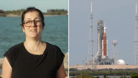 A BBC journalist speaks to camera in a black top, with the a spacecraft in position on the ground on the right.