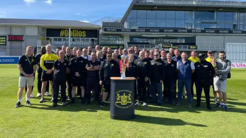 Gloucestershire CCC Staff and players of Gloucestershire County Cricket Club stand for a team shot on the pitch at the County Ground in Bristol behind a plinth on which rests the Vitality Blast T20 trophy which they won at finals day at Edgbaston in Birmingham. It is a sunny day and the pavilion is visible in the background.