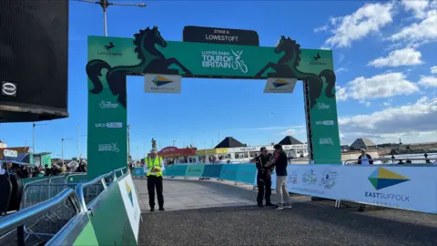 Richard Knights/BBC A green Lloyds Bank Tour of Britain banner over a paved surface with Lowestoft's South Pier visible behind. There is a blue sky with white clouds and several spectators and race officials standing around.