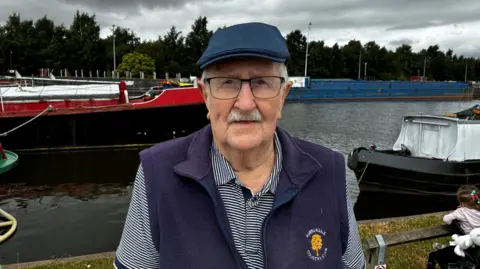 Brian wears a blue flat cap, striped top and a pair of glasses. He stands at the side of the water and a red barge is behind him.