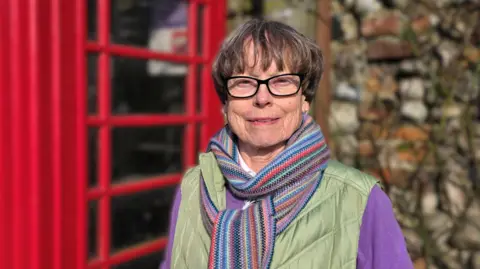 Paul Moseley/BBC A woman wearing glasses, a green bodywarmer, and a multicolored scarf stands in front of a bright red telephone booth. The background includes a stone wall and soft-focus surroundings. 