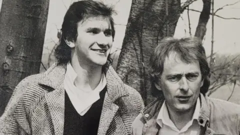 Handout A black and white photo shows a taller man with dark shoulder length hair who is smiling and wearing a shirt, jumper and coat and a man with hair which is shoulder length at the back who is wearing a shirt and jacket. They are standing in front of two tree trunks.