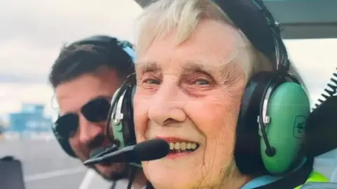 Joan Airey is sitting in the front seat of a helicopter and smiling brightly at the camera. She has short, white hair and is wearing a large green headset. A man is poking his head out from behind her.
