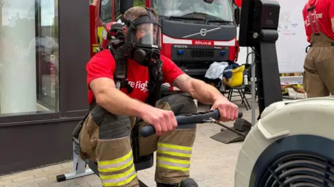 A man is exercising on a rowing machine. He has an oxygen mask and visor over his face with a red tank strapped to his back. He is wearing a red T-shirt and the trousers from a firefighting uniform. 
