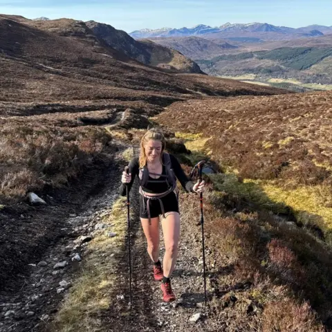 Laura Baillie Person hiking uphill on a narrow mountain trail using trekking poles, surrounded by rolling heather-covered hills and distant snow-capped peaks under a clear blue sky.