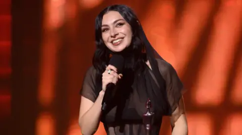 Getty Images Charli XCX smiling while accepting one of her five Brit awards on stage last year. She is wearing a sheer black dress.