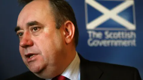 Alex Salmond in a dark suit, with a Scottish government sign and saltire in the background