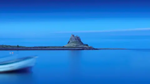Arquivo histórico da Inglaterra Um trecho plano de ilha com um castelo com torres em um afloramento solitário pode ser visto à distância, com uma extensão de água azul parada em primeiro plano que se mistura com o céu azul escuro. A borda de um barco azul e branco em primeiro plano está fora de foco.