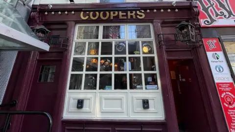 Coopers pub front has a cream painted central window and two doors either side. The frontag eis painted purple and the name Coopers in written in gold.