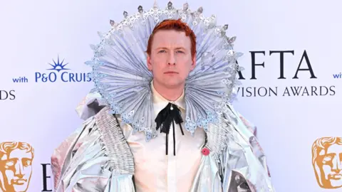 A man with a shiny outfit on looking into the camera and smiling. He has red hair and he has a metallic ring around his face/ head. 