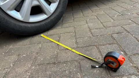 Martin Heath/BBC Car tyre in the top left corner, parked on block paving. A yellow tape measure in an orange and black sleeve is measuring the distance between the tyre and the kerb.
