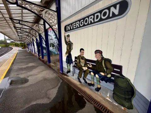 David May A mural of three men dressed in traditional Scottish wear sitting on a bench in Invergordon train station. An INVERGORDON sign is above the mural.