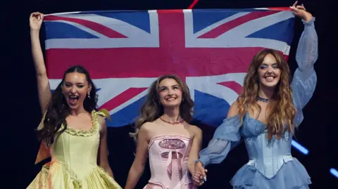 Reuters Three young women walking together and holding hands, wearing bright costume dresses holding a union jack flag above their heads