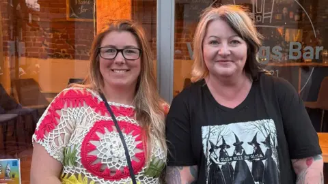 Lauren Emms A woman with long light brown hair stands in front of a pub alongside another woman with short blonde hair with some dark sections. The woman on the left has glasses and wears knitted top. The woman on the right wears a black T-shirt. 