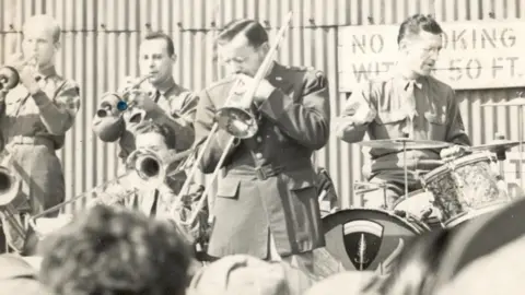 Glenn Miller Collections/University of Colorado Boulder A black and white image of the band, showing four musicians, all wearing military uniform. Drummer Ray McKinley is on the right with a small drumkit. To his left is Glenn Miller on the trombone.
